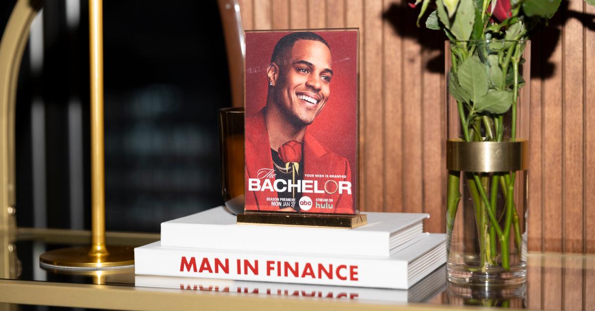 A photo card of 'The Bachelor' star Grant Ellis sits on top of white books. The spine of the bottom book says "Man in Finance" in red coloring.