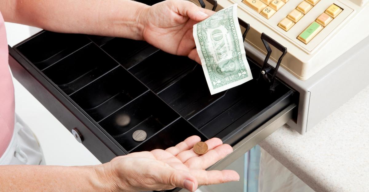 A person holds a dollar and a penny in front of an open cash register