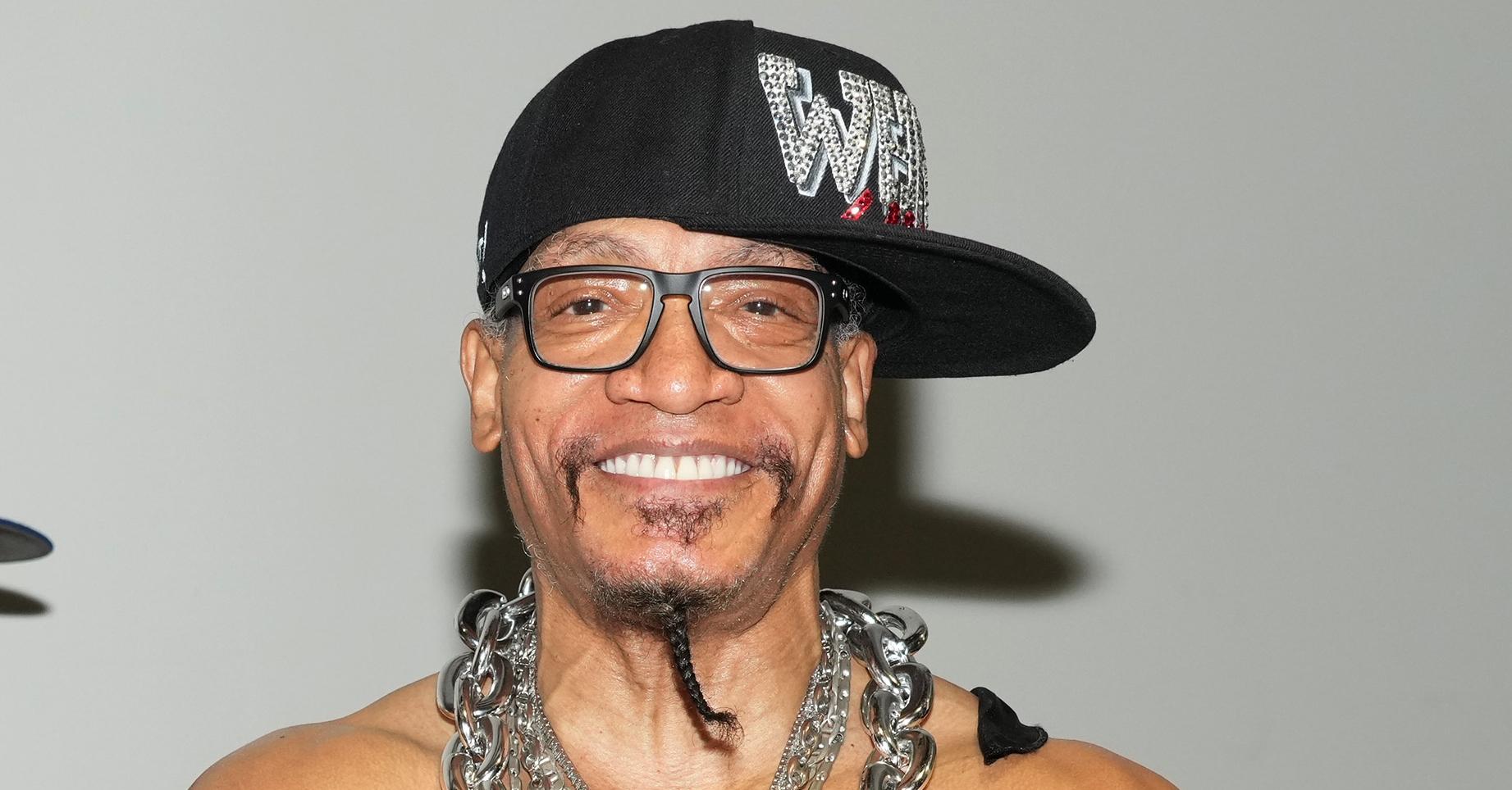 Melle Mel smiles with his shirt off wearing large chains, glasses, and a bedazzled hat.