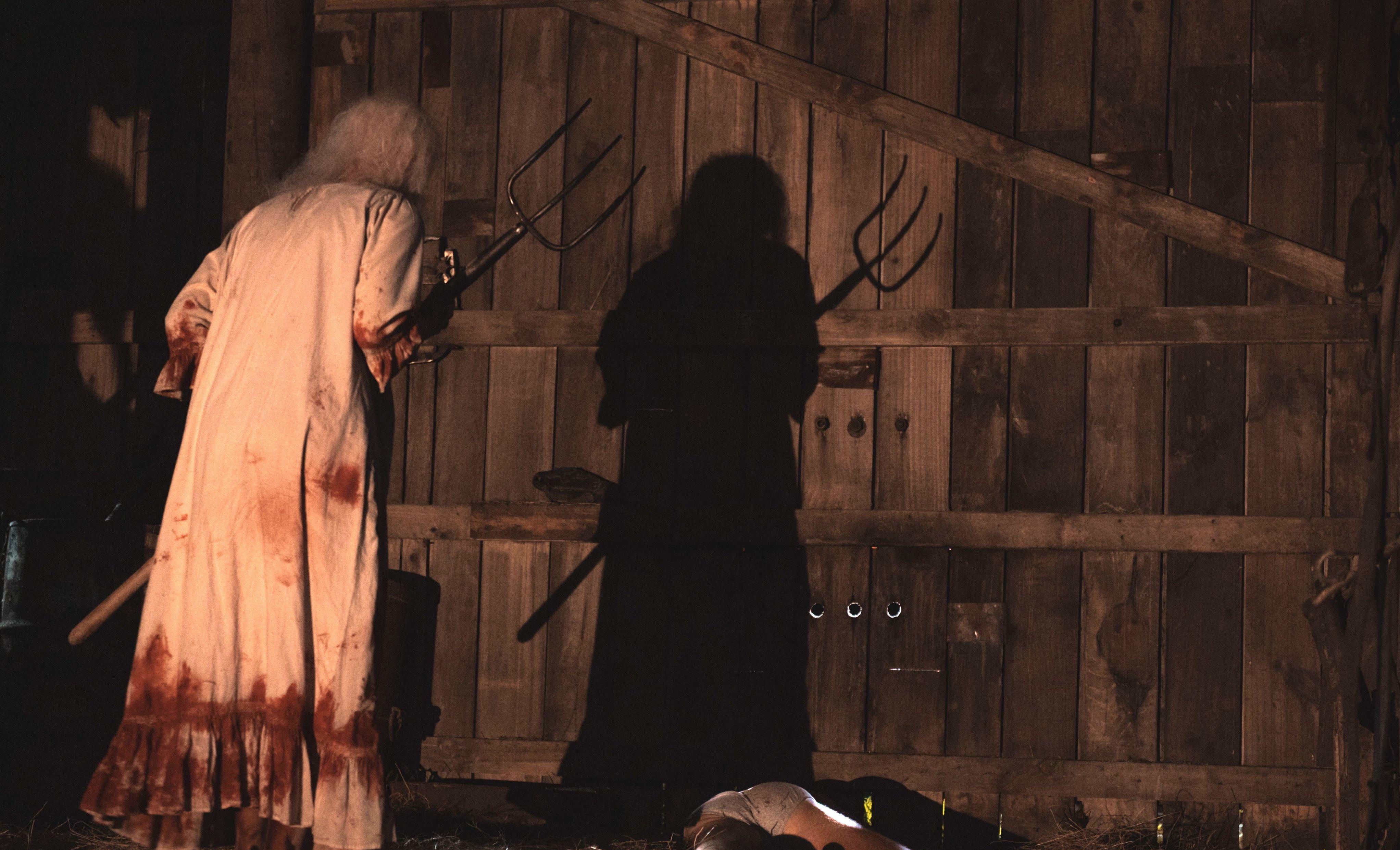 An elderly woman is wearing a blood-soaked nightgown while wielding a pitchfork in 'X.'