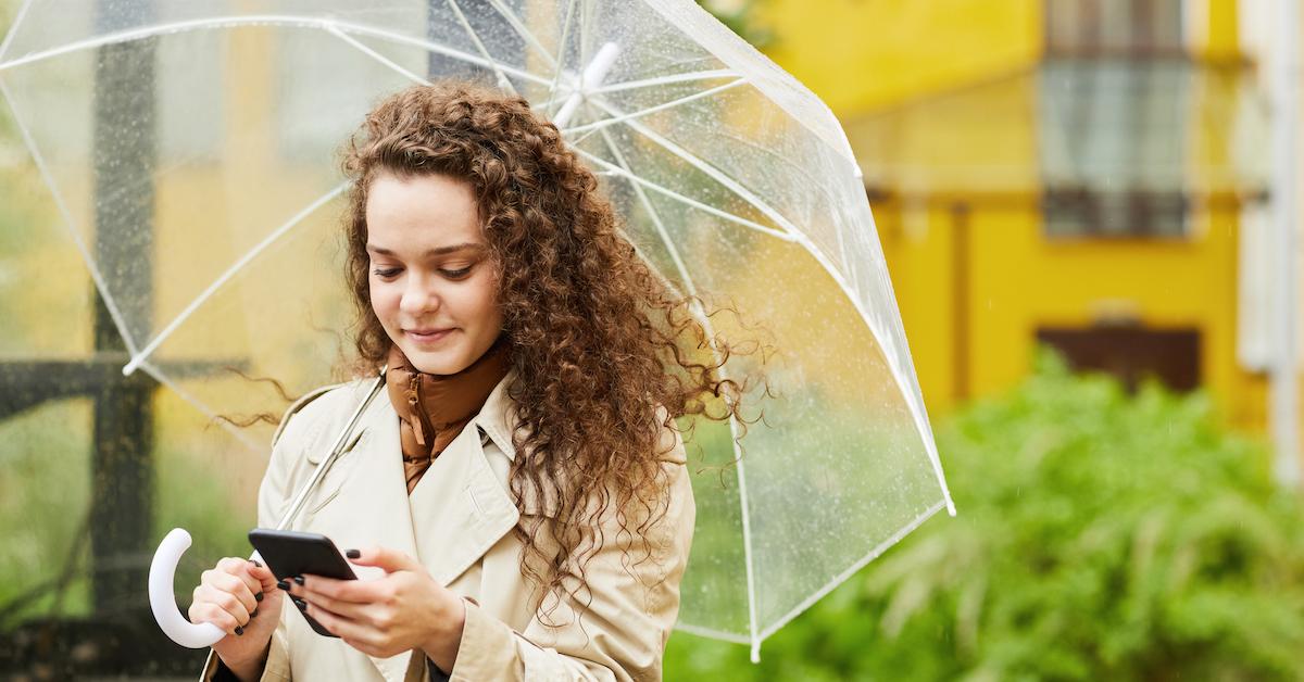 woman with curly hair stands under umbrella and wears trench coat, looking at her phone