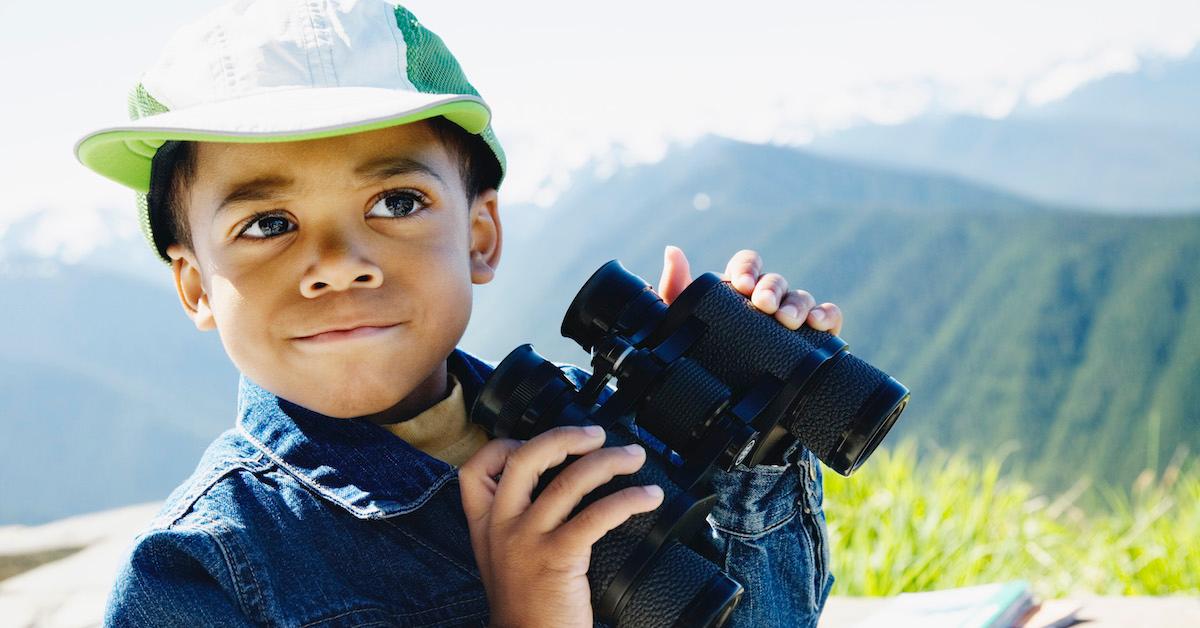 A little boy wearing a hat and holding binoculars with a mountain in the background