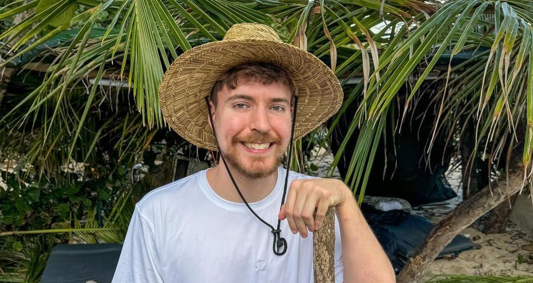 MrBeast smiling while wearing a floppy hat in front of palm trees