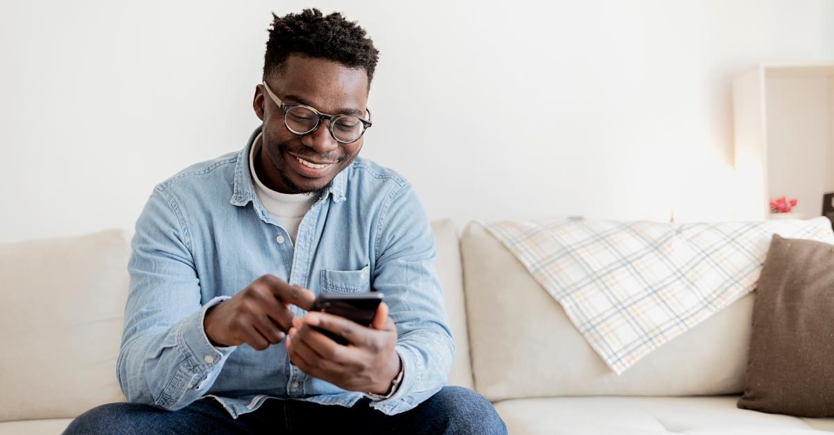 Stock image of a Black man in a blue shirt smiling as he scrolls on his cell phone 