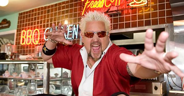 Guy Fieri, in a red and white shirt, strikes a pose inside a diner