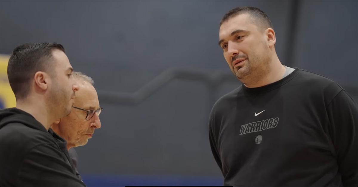 Dejan Milojević talking with other coaches in a Warriors sweatshirt. 