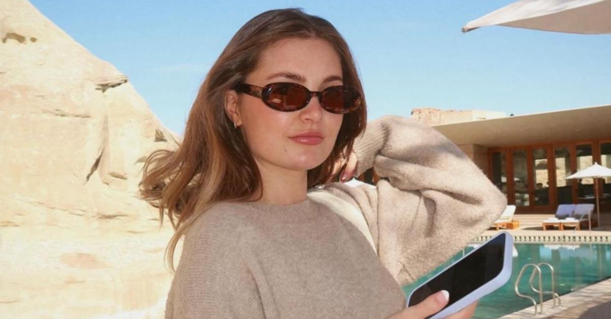 Aspyn Ovard in sunglasses and sweater while holding phone outside by a pool