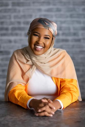 Muna wears an orange hijab and white t-shirt, smiling for her official portrait for Season 2 of 'The Mole.'