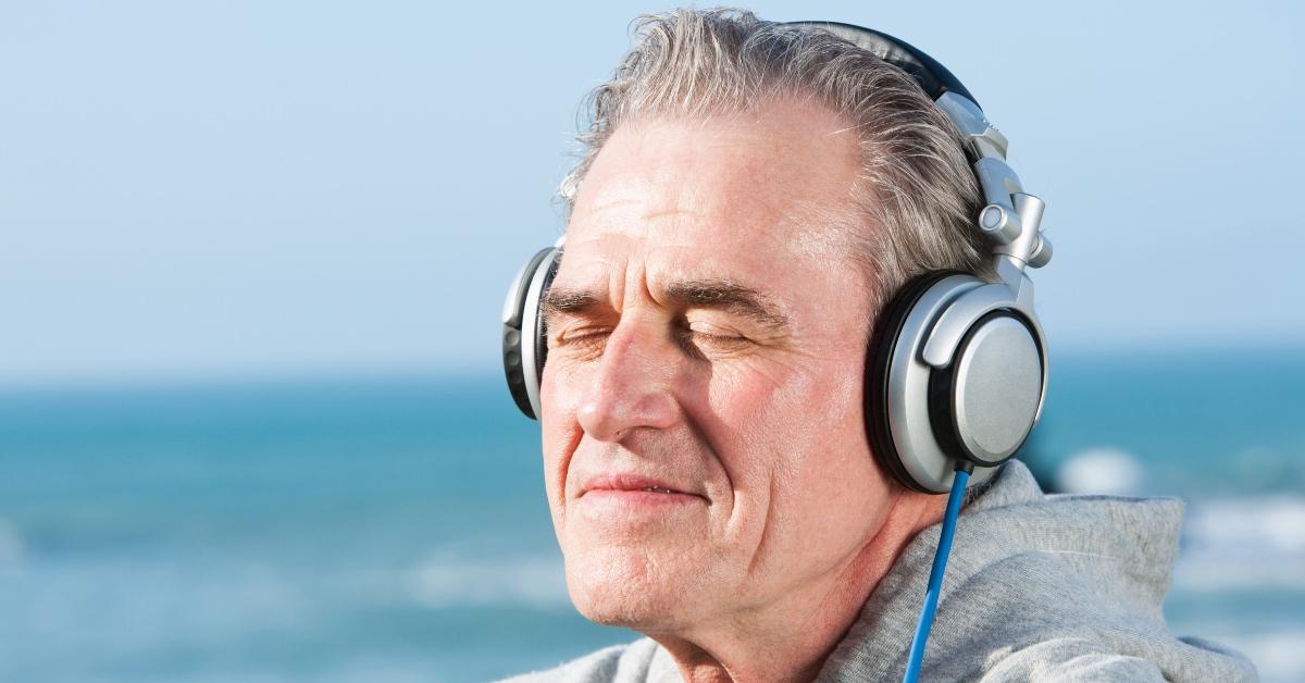 An older man wears noise canceling headphones with his eyes closed by the sea.