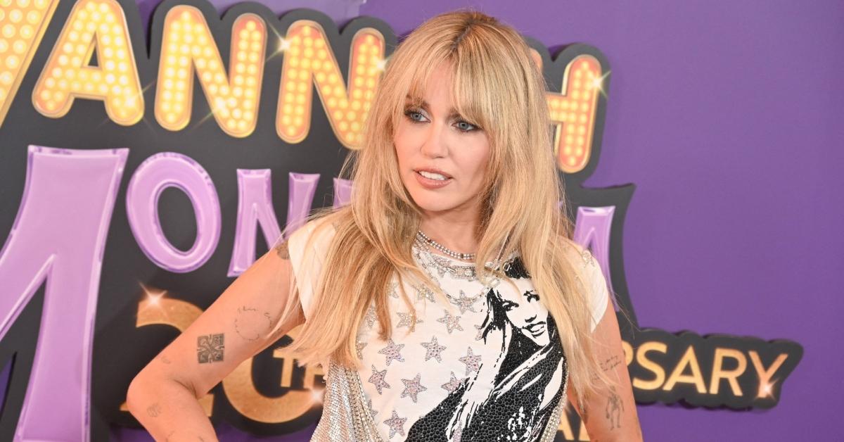 Miley Cyrus wears a Hannah Montana shirt in front of the Hannah Montana Anniversary banner.