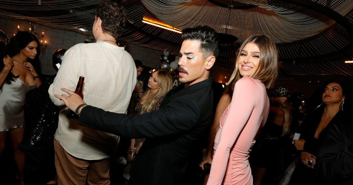 Tom Sandoval looking down at a party as Rachel Leviss poses over her shoulder up against his back