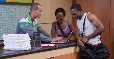 Gym employee types on a laptop as two gym clients look on.