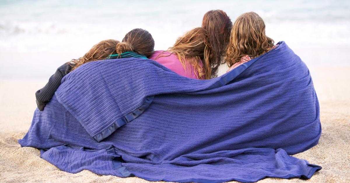 Rear view of a group of friends sitting on the beach wrapped together in a blanket - stock photo