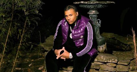 Fanum sits next to a fountain and looks serious wearing a purple and black tracksuit and purple vest.