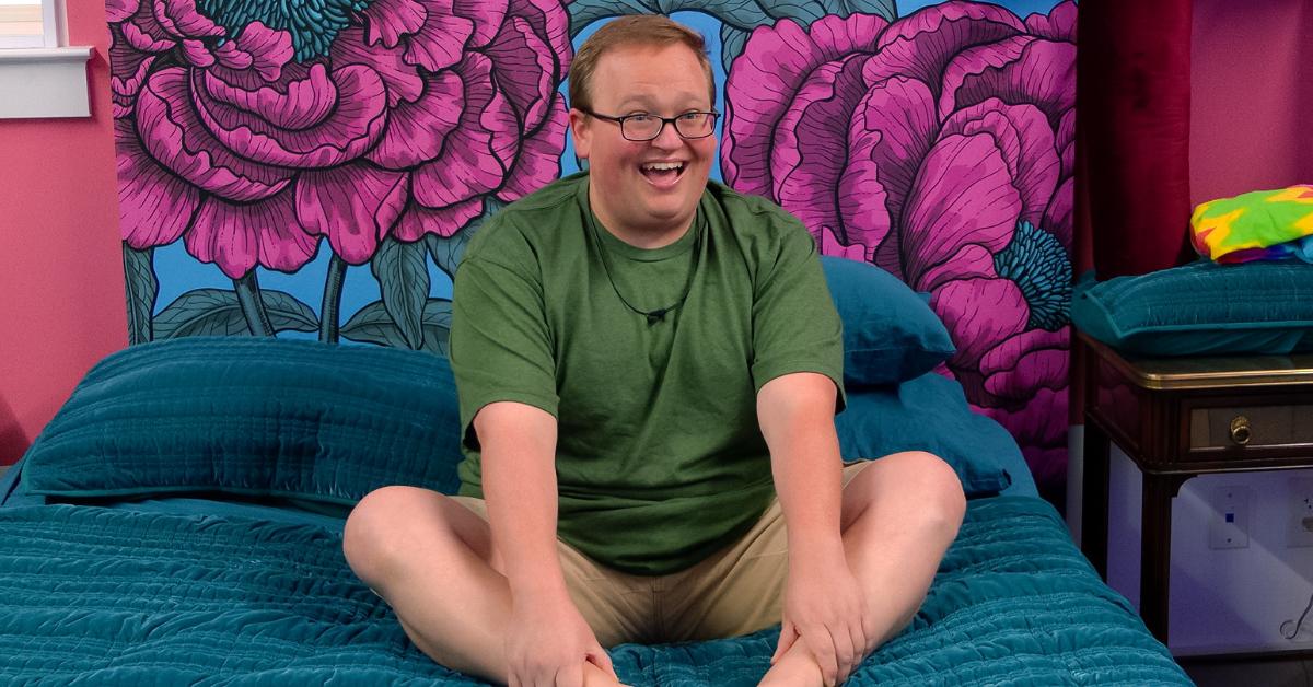 Brandon, u kaki kratkim hlačama i zelenoj majici, smiješi se dok sjedi na svom krevetu u 6. sezoni 'The Circle'.