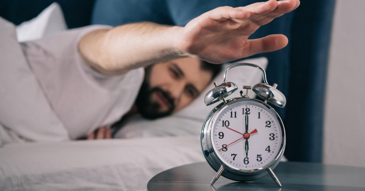 bearded young man trying to turn off alarm clock while lying in bed picture id