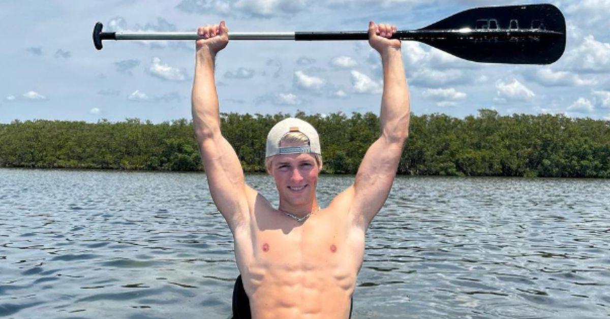 Micah Plath holds a paddle over his head while canoeing shirtless in a lake