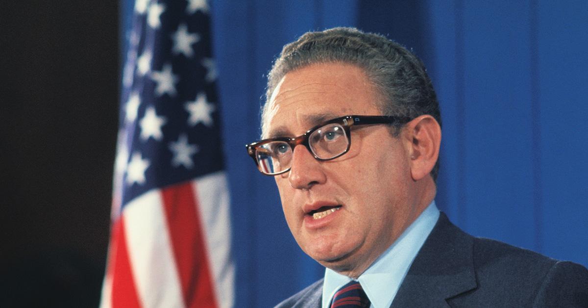 Henry Kissinger speaking with a flag behind him. 