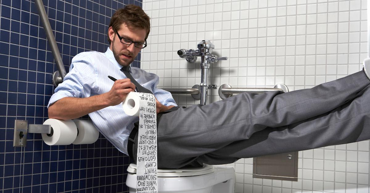 Businessman sitting on toilet with feet up, writing list on paper - stock photo