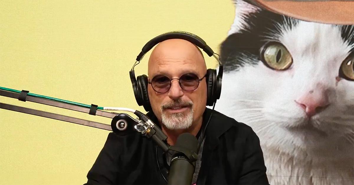 Howie Mandel speaking into a mic with a cat behind him. 