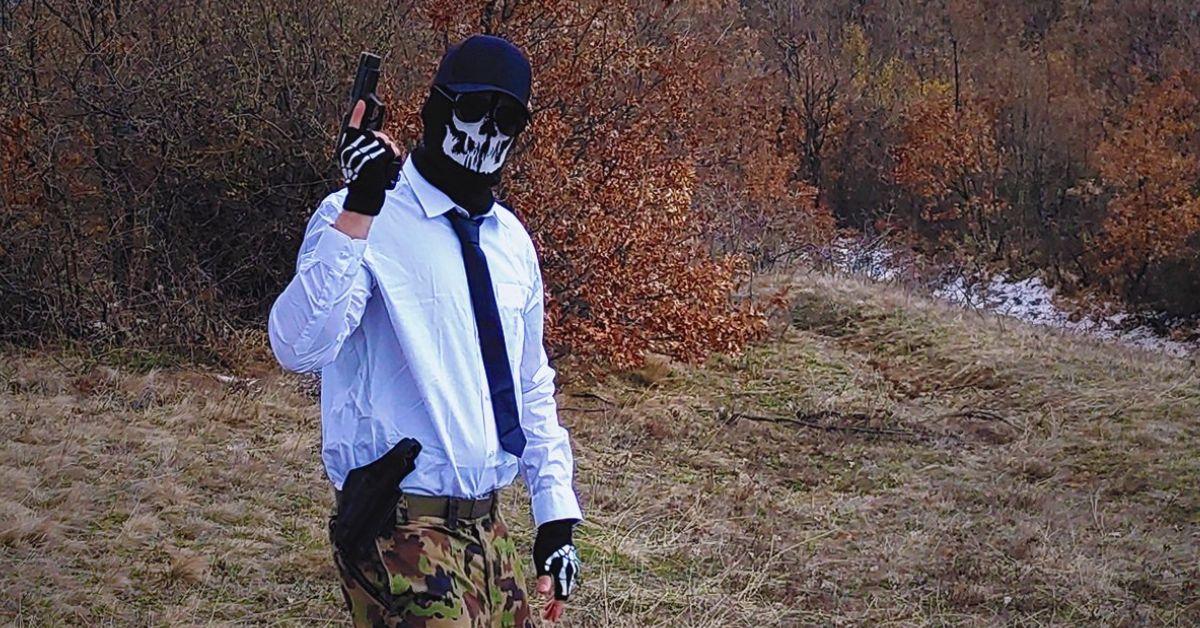 Shadman poses outside with a gun wearing a skeleton mask, gloves, camo pants, and a button down shirt with a tie