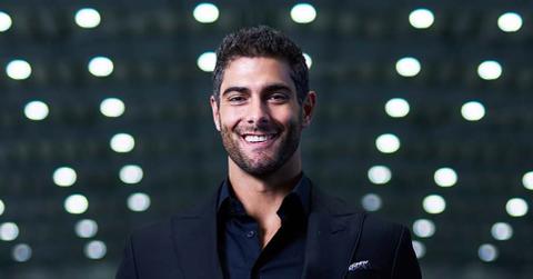 Jimmy Garopollo smiles in football stadium wearing a suit and holding a raiders jersey.