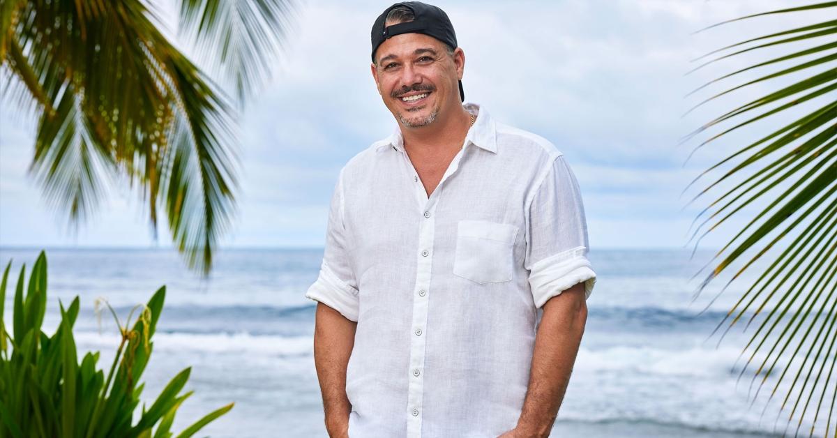 Boston Rob stands on a beach surrounded by palm trees