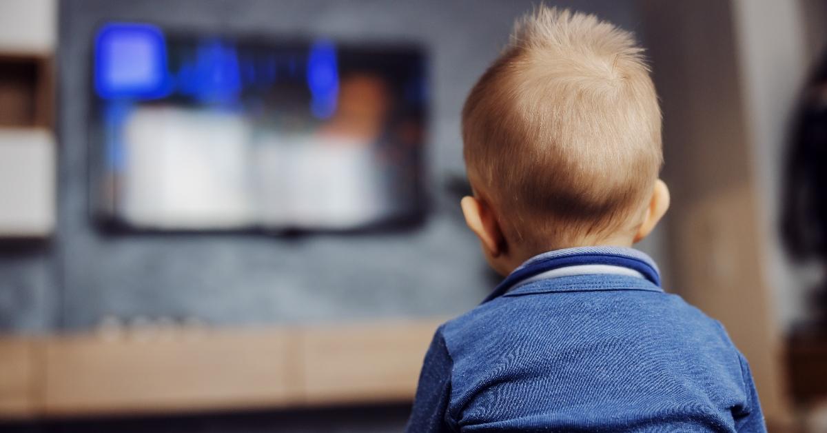 Rear view of focused blond little boy sitting on the floor in living room and watching cartoons on television. - stock photo