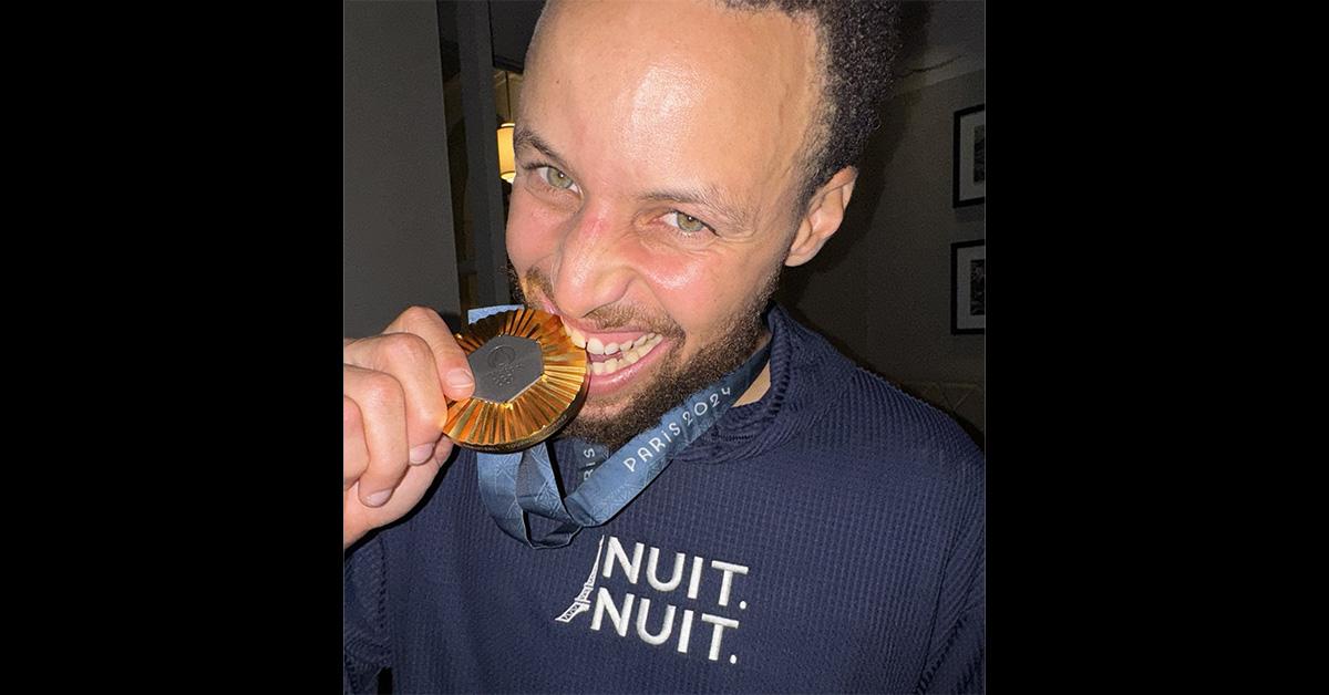 Steph Curry wearing a "NUIT. NUIT." shirt on and a gold medal. 