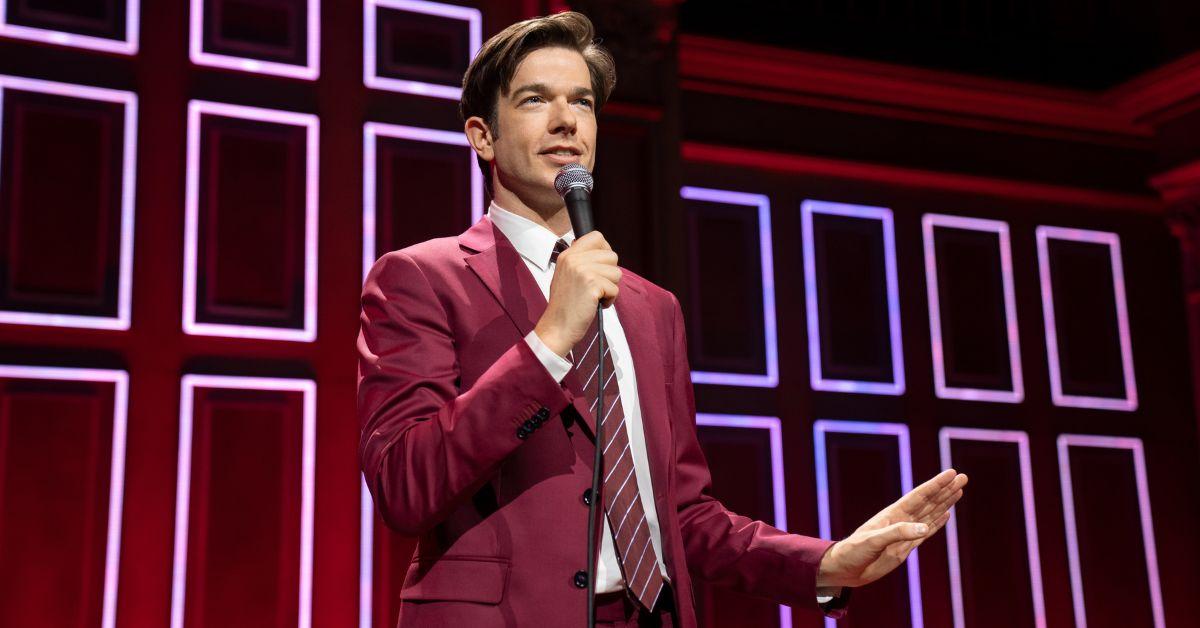 John Mulaney speaking onstage while wearing a burgundy suit and tie.
