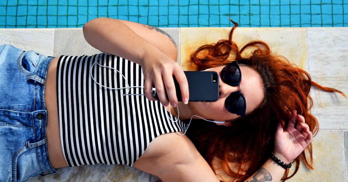 A girl lays by the pool with sunglasses scrolling on smartphone. 