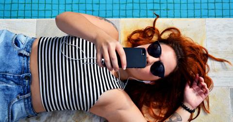 A girl lays by the pool with sunglasses scrolling on smartphone.