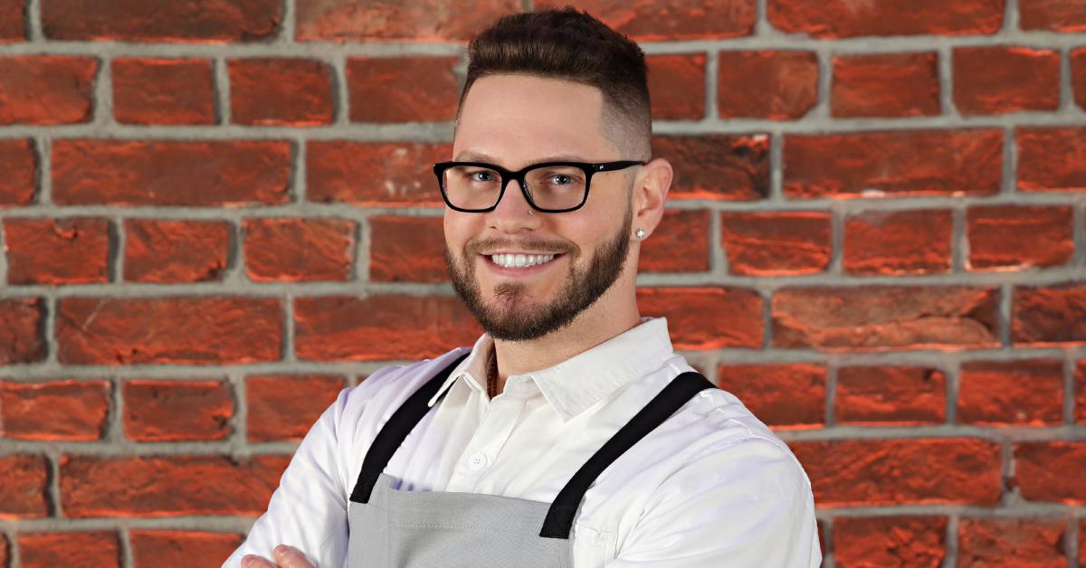 Jordan from Season 3 of 'Next Level Chef' stands in front of a brick wall wearing a gray apron.