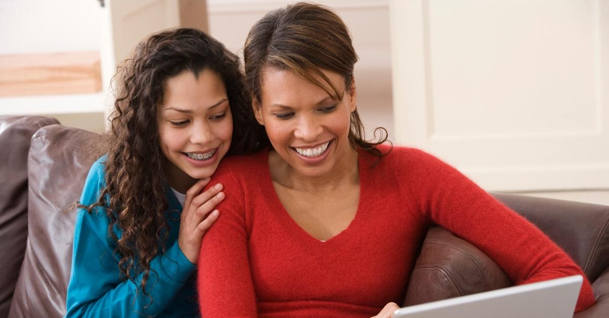 Black woman and her teenage daughter sit on the couch and scroll the internet on the computer.