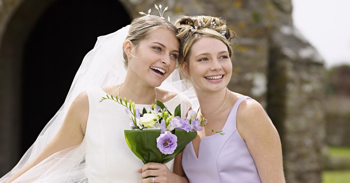 A bride wears a veil and a bouquet smiles next to her happy bridesmaid that's wearing a purple dress.