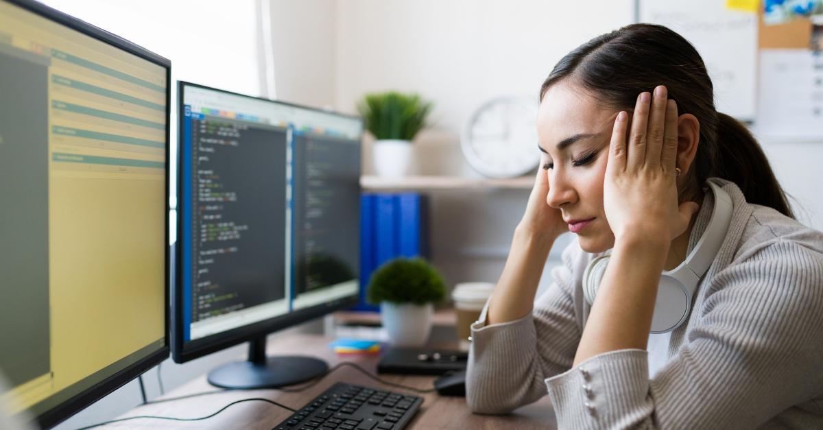 Woman looks depressed at work in front of two computer screens.