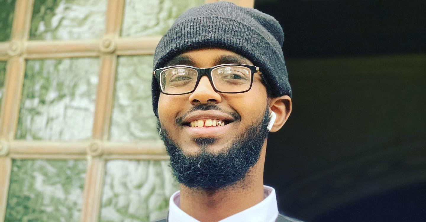 Darkest Man smiles next to a window wearing glasses and a button down shirt and bowtie. 