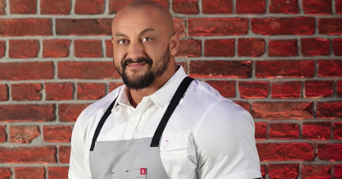 Mada from Season 3 of 'Next Level Chef' stands in front of a brick wall wearing a gray apron