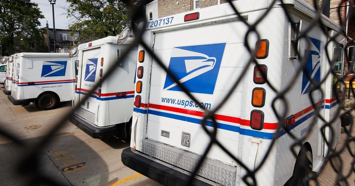 USPS trucks park in front of a barbed wire gate,