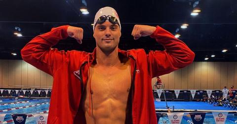 Matt Klotz wears white swimming cap and red jacket and flexes in front of competitive pool.