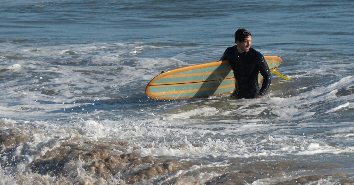 Joey Graziadei, clad in a wet suit, gazes to the side while he gracefully emerges from the ocean, surfboard in hand.