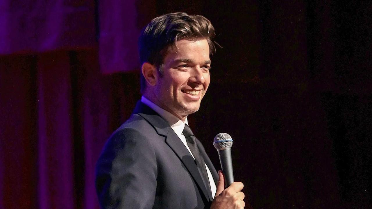 John Mulaney in a black suit performing on stage during his 'From Scratch' Tour