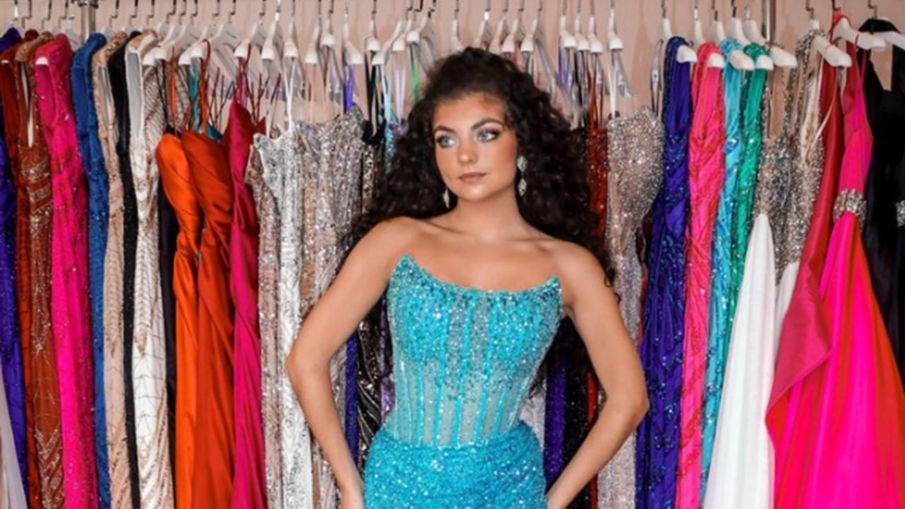 Gabriella wearing a blue gown and standing in front of a rack of gowns