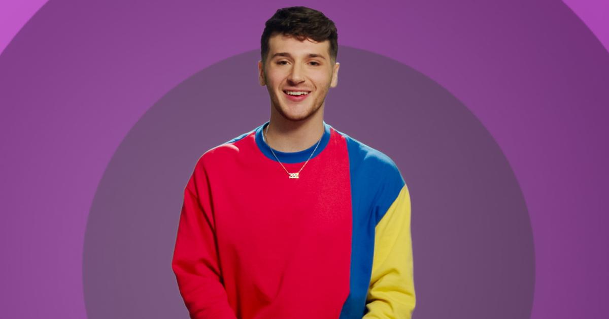 Jordan smiles and poses in front of a purple background while wearing a red, blue, and yellow long-sleeve for 'The Circle' Season 6.