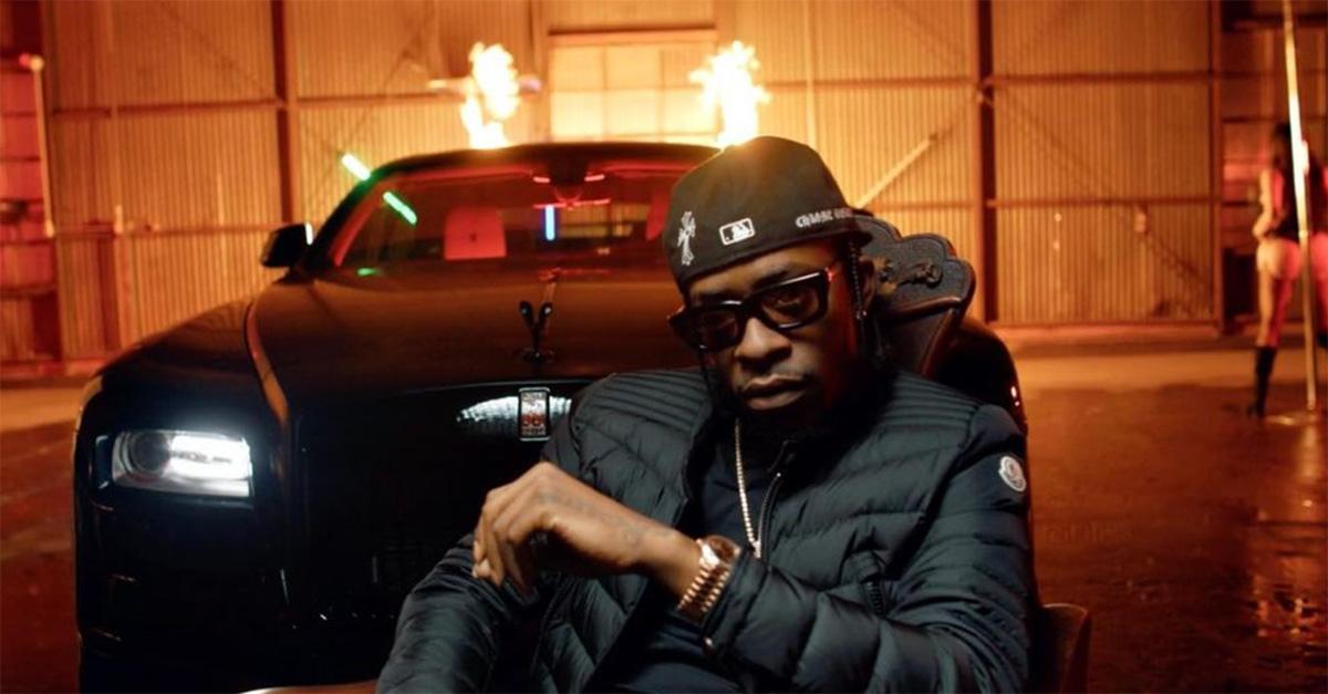 Rich Homie Quan in front of a car with flames in the background.