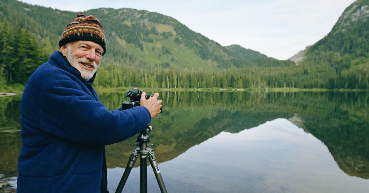 Senior man setting up camera by lake, looking over shoulder