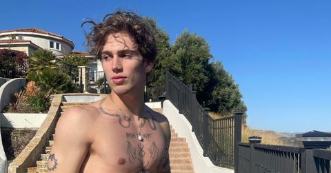 Vinnie Hacker has his shirt off and looks over his shoulder while standing outside of a mansion.