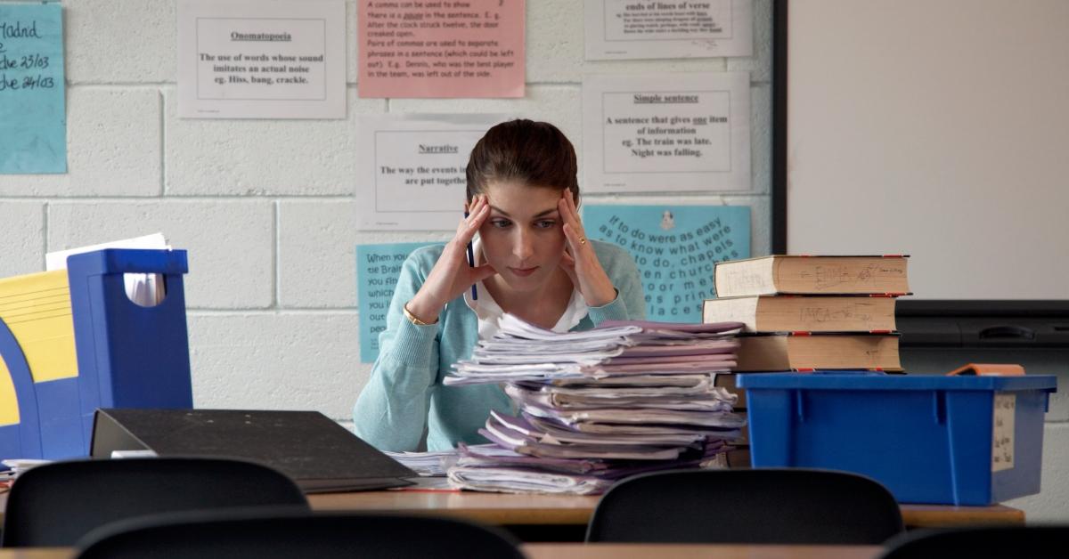 Schoolteacher at desk staring at piled exercise books, hands to head stock photo