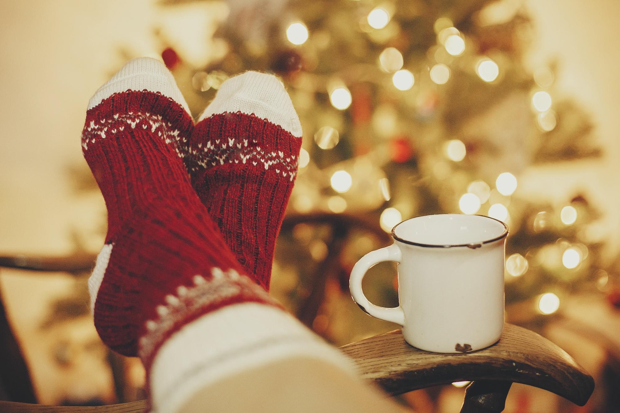 festive socks and mug with hot drink on old wooden chair on background of golden beautiful christmas tree with lights in festive room. cozy winter holidays. warm atmospheric moment