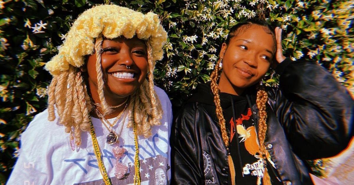 Bobbi Lanea and Folayan Kunerde have braids and wear t-shirts and smile and pose outside near a plant wall with white flours.